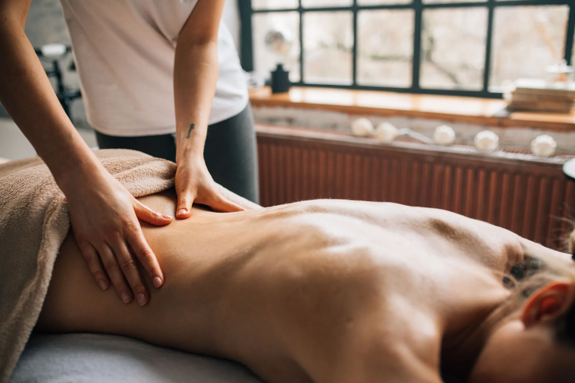 A massage therapist's hands working along a guest's back during an outcall massage, next to a bright window